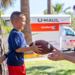 A Moving Helper hands a football to a small child as the child’s mother stands behind her son.