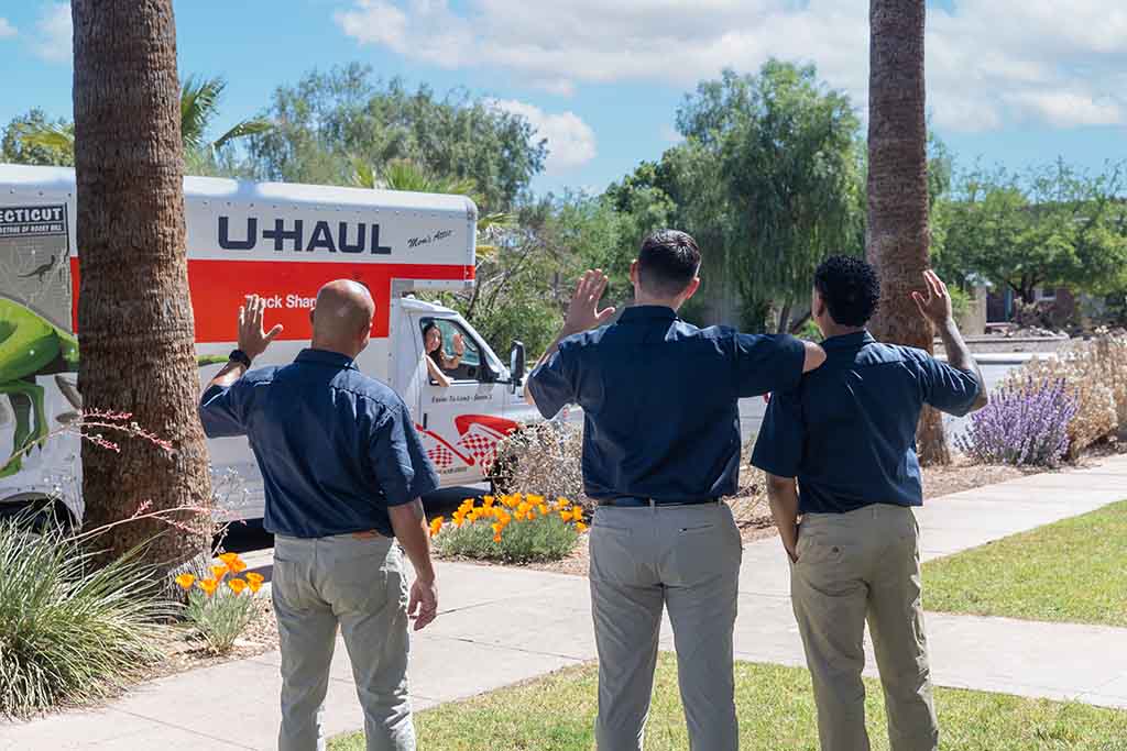 Three Moving Helpers wave goodbye to a female customer who is riding in the passenger seat of a U-Haul truck rental that is driving away.